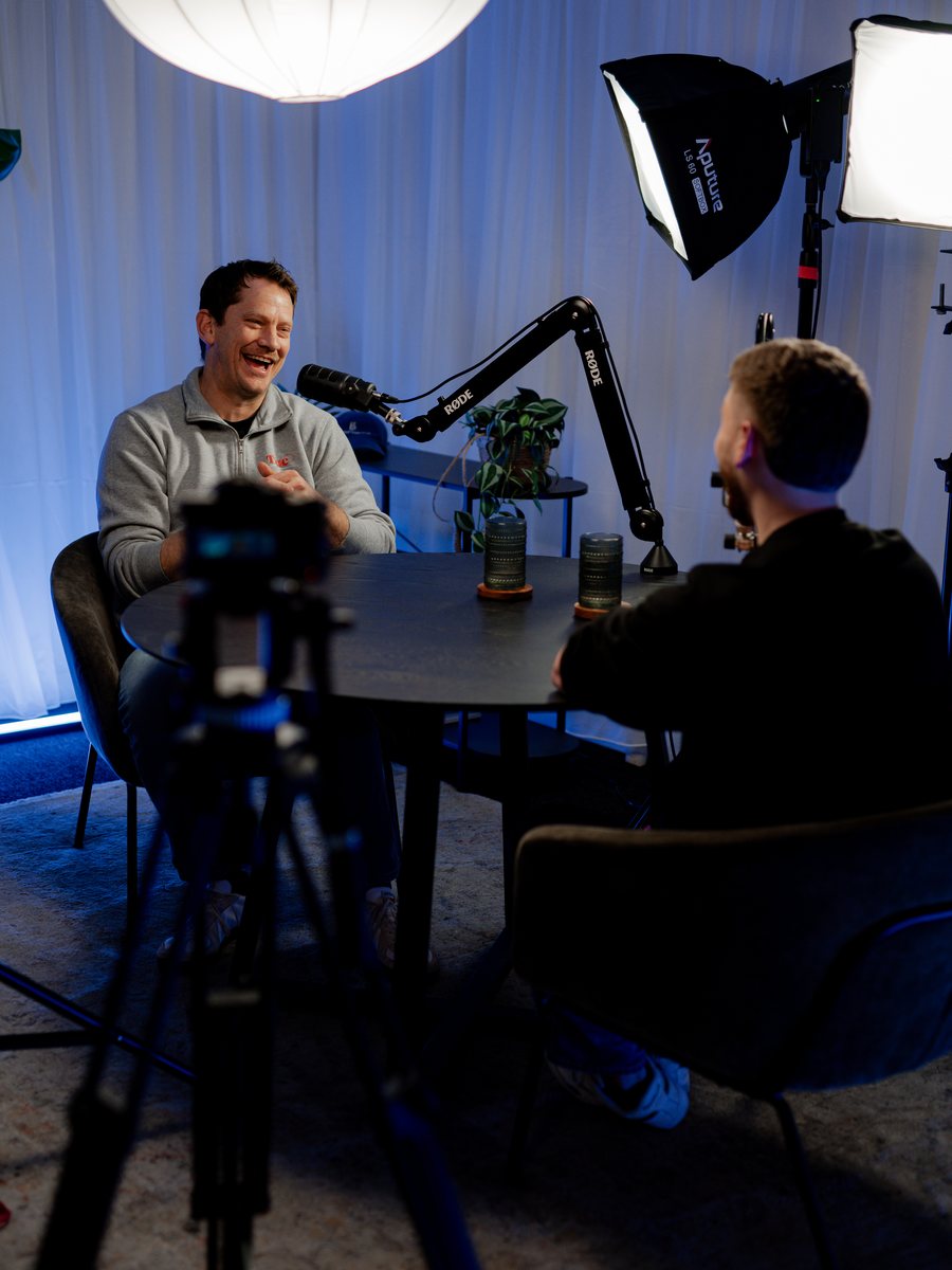 The Better Conversations studio mid recording: Stu Moerman talking across a dark table, microphones on booms between him and the host, a camera in the foreground.